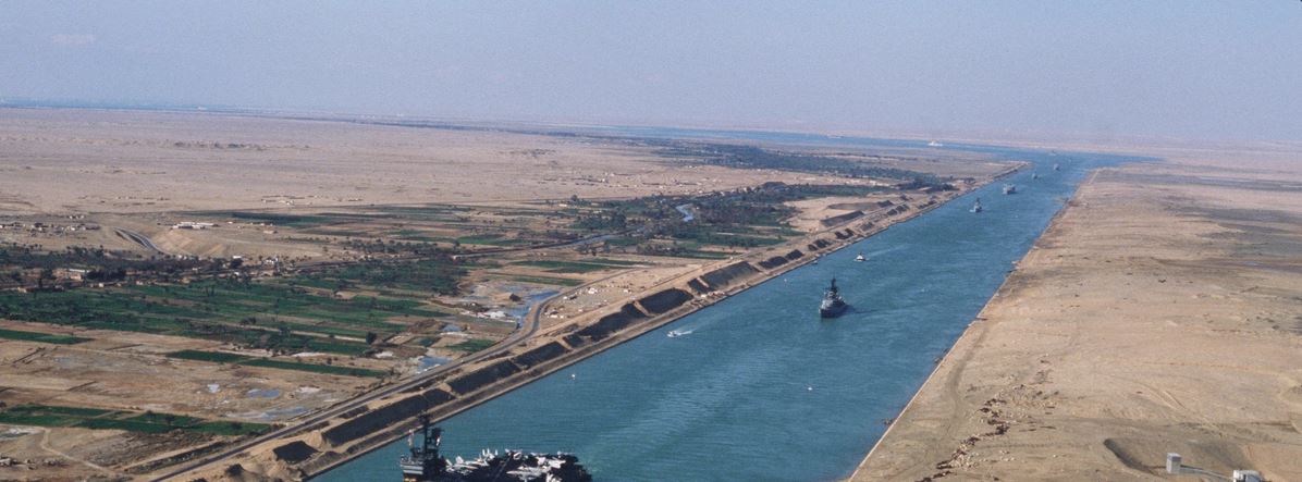 Aerial view along the Suez Canal, a straight blue channel between desert banks with an aircraft carrier and escort ships in transit